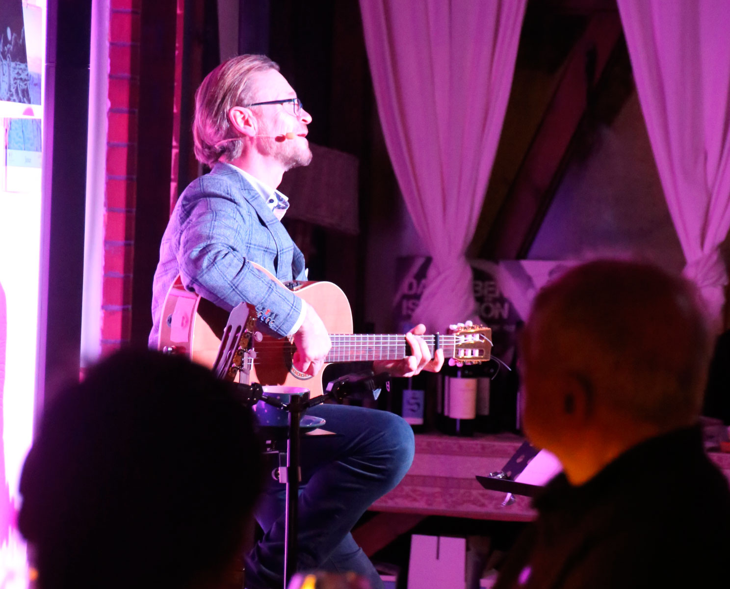 Mann sitzt auf einem Hocker und spielt Gitarre im Vordergrund des Bildes sitzen Menschen und schauen ihn an. 